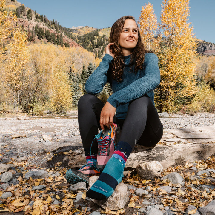 Woman sitting by a riverbed with one shoe off, wearing teal mountain micro crew socks