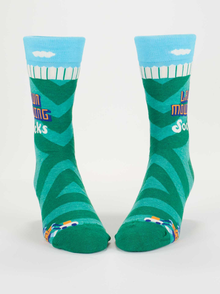Blue Q Lawn Mowing Socks M-Crew Socks feature a green grass pattern, blue sky with clouds at the top, "Lawn Mowing Socks" on the sides, and small lawn mower graphics near the toes. Fits most men&