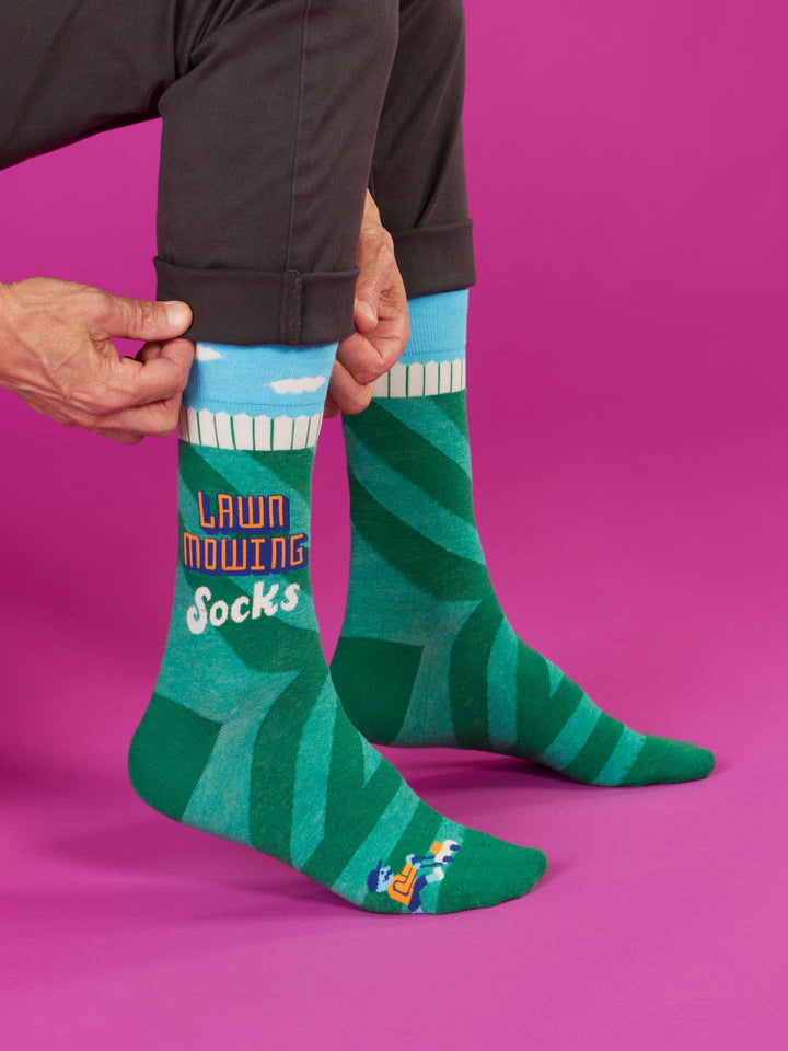 A person reveals Blue Q Lawn Mowing Socks M-Crew Socks, made of colorful combed cotton with a green lawn print and "Lawn Mowing Socks" text on a pink background, perfect for any men&