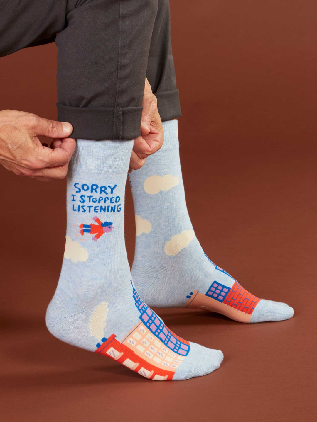A person pulls up Blue Q Sorry I Stopped Listening M-Crew Socks—light blue combed cotton, men’s size 7-12, with clouds, buildings, a flying superhero, and “SORRY I STOPPED LISTENING” in blue and orange lettering.
