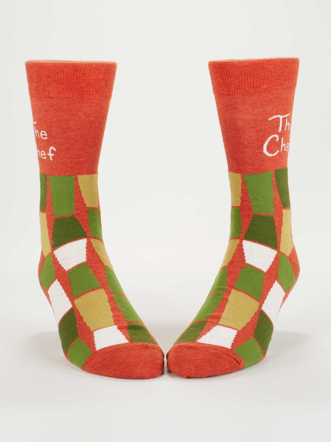 The Blue Q "The Chef" M-Crew Socks feature red fabric with green, yellow, and white geometric patterns, "The Chef" in white text at the top, fit men’s shoe size 7-12, and are made from soft combed cotton. Displayed upright on a plain white background.
