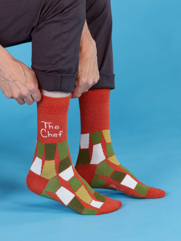A person wears Blue Q The Chef M-Crew Socks—orange, green, and white patterned combed cotton socks with "The Chef" on one, sized for men&