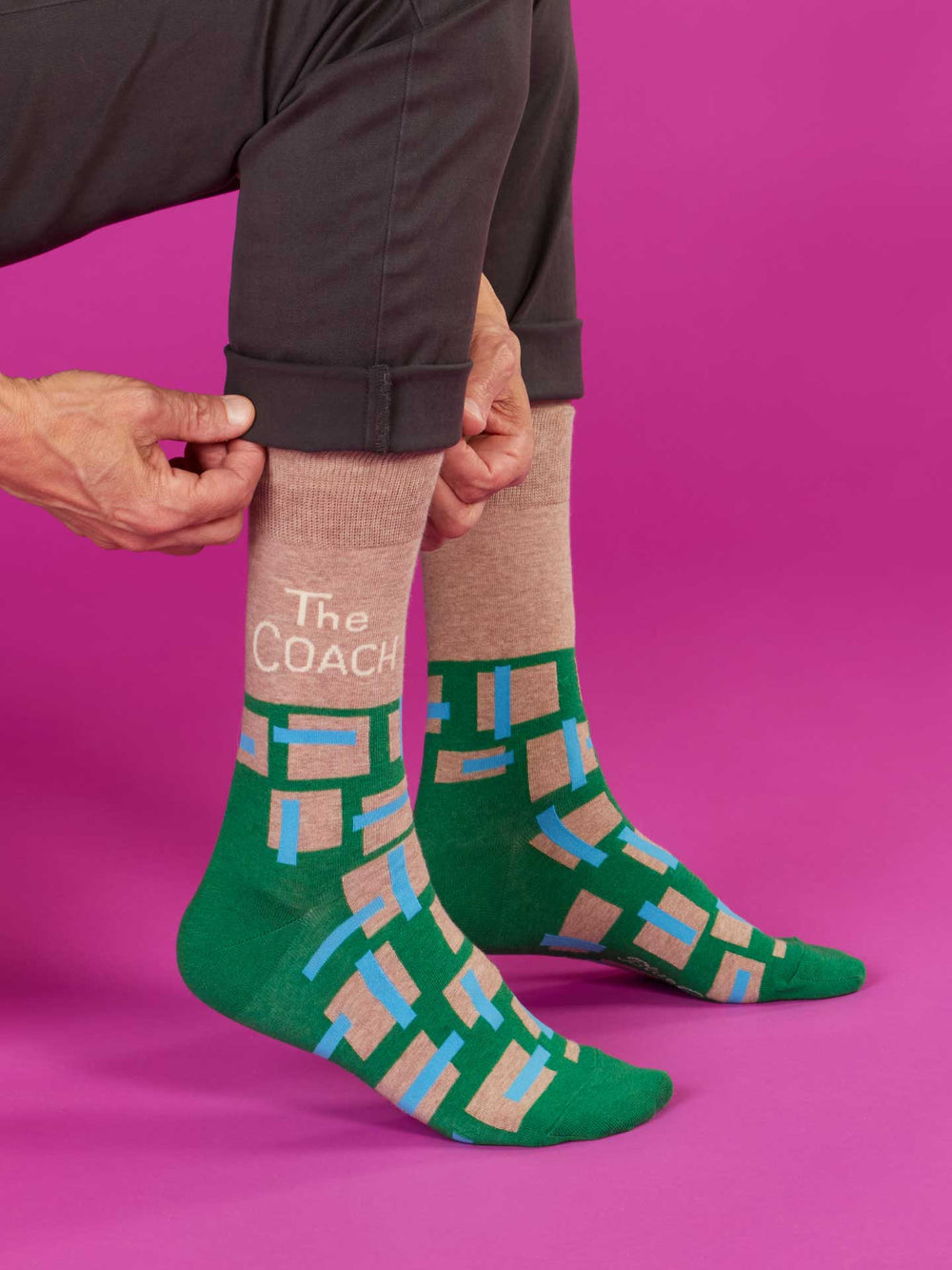 Against a pink background, a person adjusts rolled-up pants to show Blue Q's The Coach M-Crew Socks—green combed cotton with tan, blue, and pink geometric designs. "The COACH" is printed on one sock; fits most men’s shoe sizes.