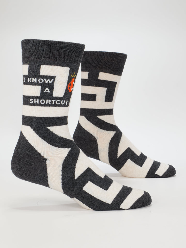 Blue Q’s I Know A Shortcut M-Crew Socks feature dark gray cotton, a white maze pattern, “I KNOW A SHORTCUT” text, and a small red ladybug graphic near the top. Fits men’s sizes 7-12.