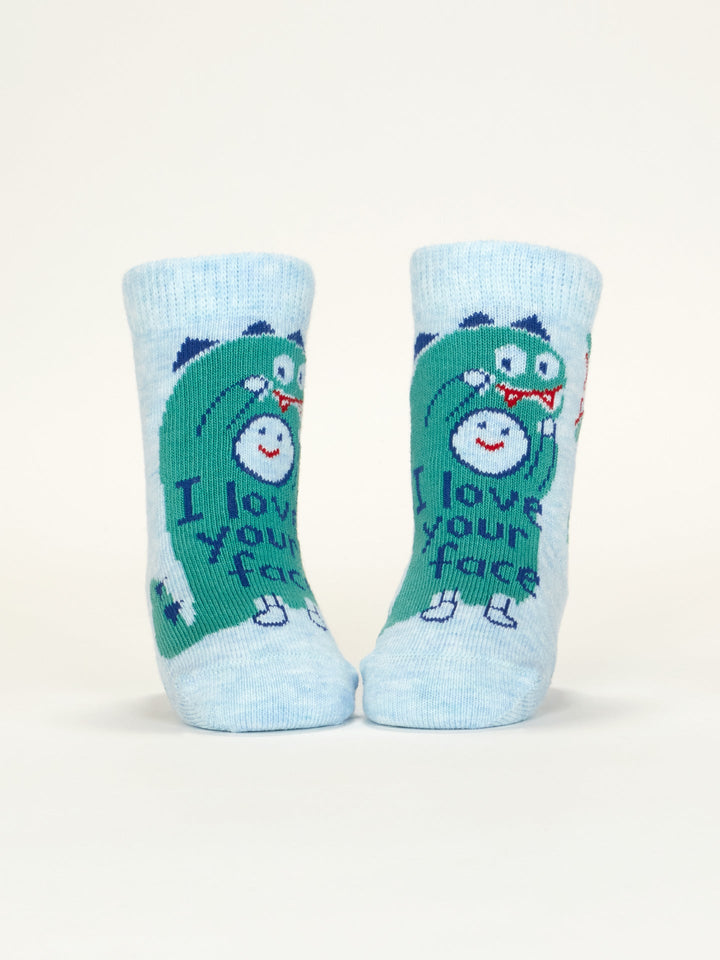 A pair of Blue Q “I Love Your Face/If You Drop Me I’ll Remember” baby socks, light blue with a green dinosaur design, made from OCS certified organic cotton. They stand upright against a plain white background.