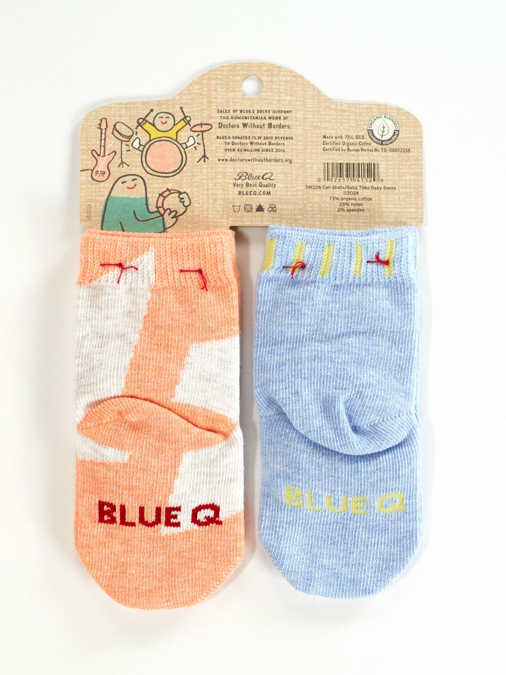 A 2-pair pack of Blue Q “I Call The Shots Now/It's Baby Time” OCS-certified ankle socks: one peach with white/red accents, one light blue with yellow/red accents. Packaging shows playful cartoon characters and the Blue Q brand.