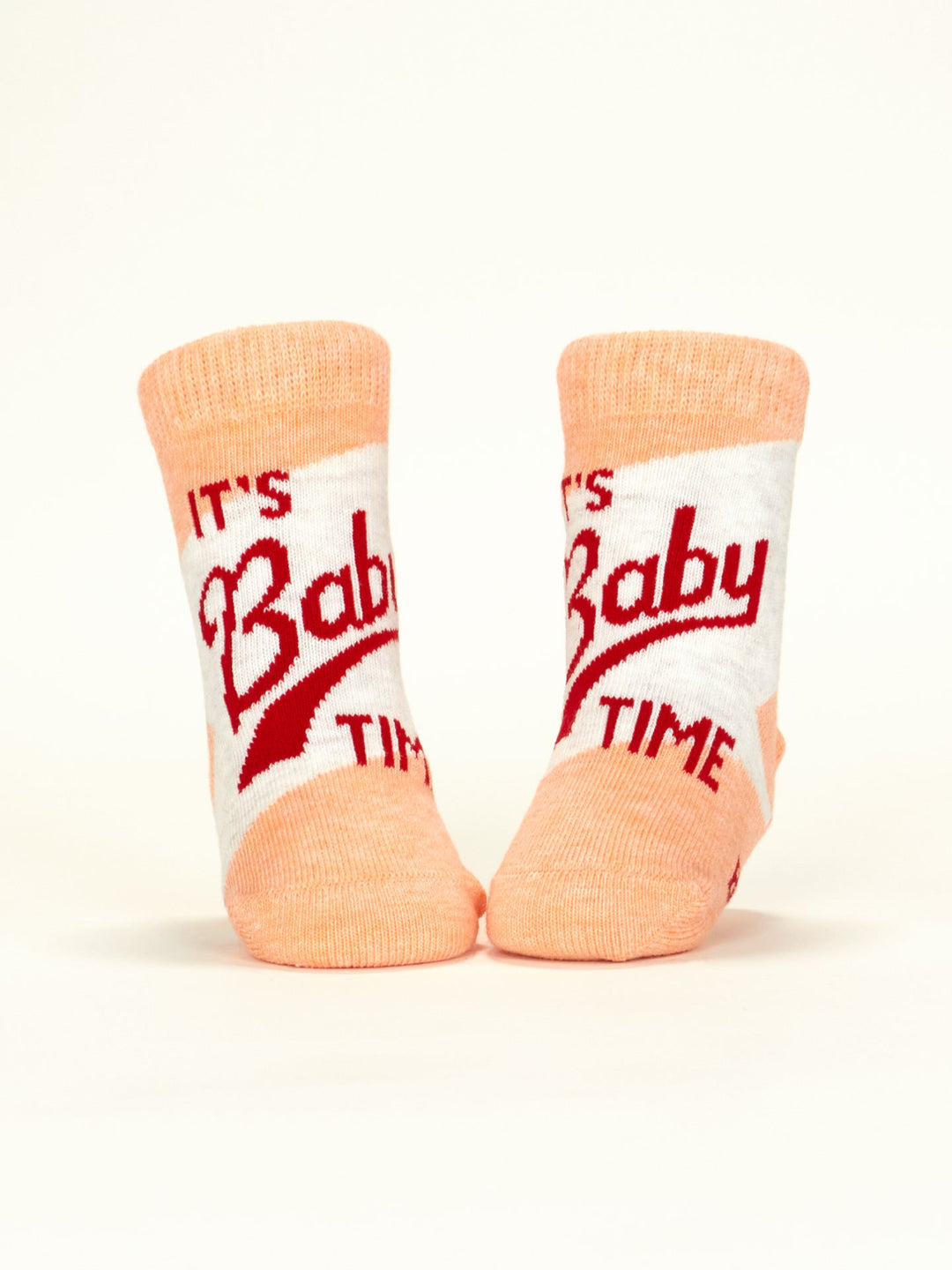 A pair of Blue Q “I Call The Shots Now/It’s Baby Time” peach and white baby socks with red text, displayed upright against a light background—perfect to pair with our cozy Family Pants or soft Organic Cotton Pants.