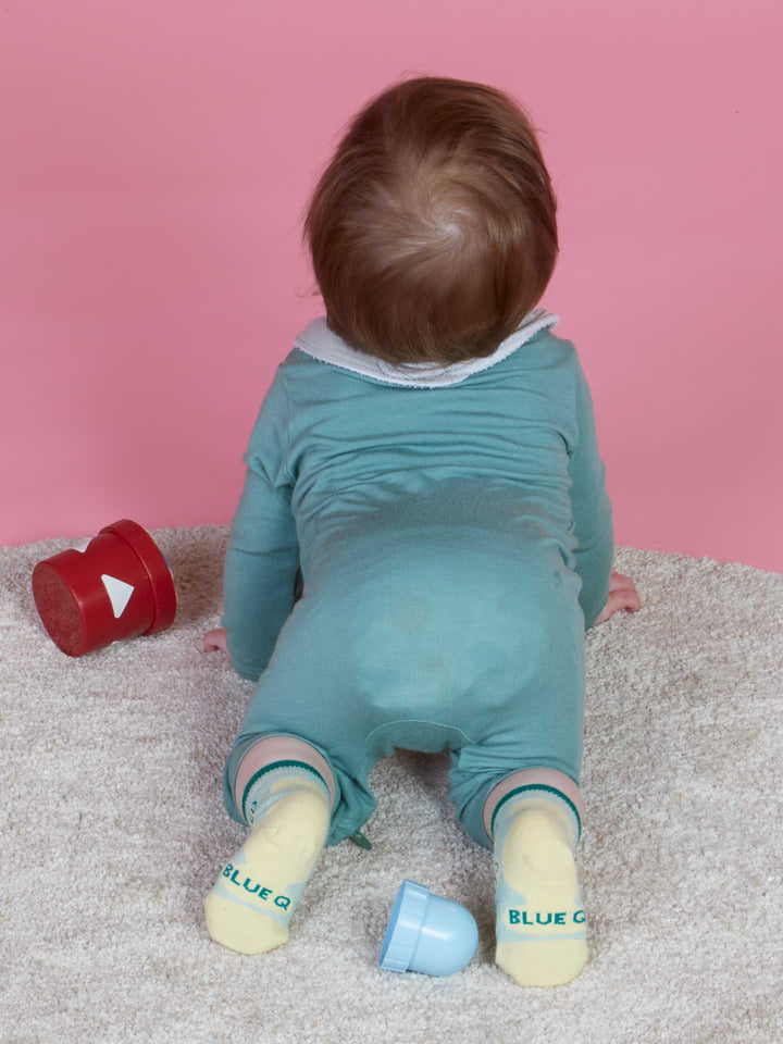 Wearing Blue Q’s Certified Cuddle Bug/I Bet I Can Make Us Both Cry Baby Socks, a baby in a teal onesie crawls on a beige carpet toward a pink background with their back to the camera; red and blue toy cups are scattered nearby.