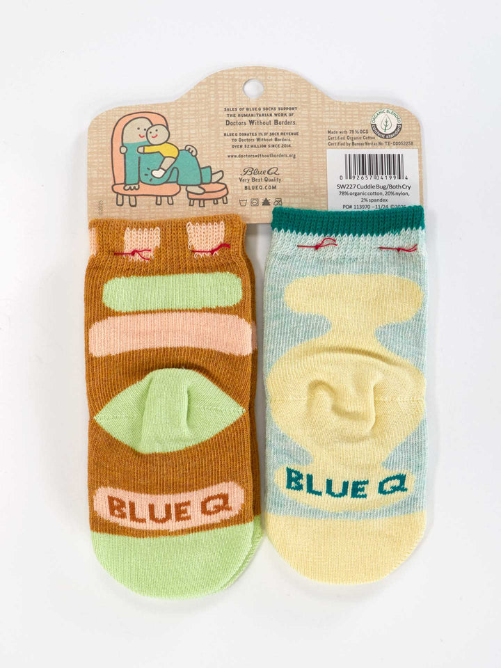 Certified Cuddle Bug/I Bet I Can Make Us Both Cry Baby Socks by Blue Q: a playful, OCS Certified pair with cartoon designs—one brown/green with animals, the other blue/yellow abstract—attached to a branded cardboard hanger.