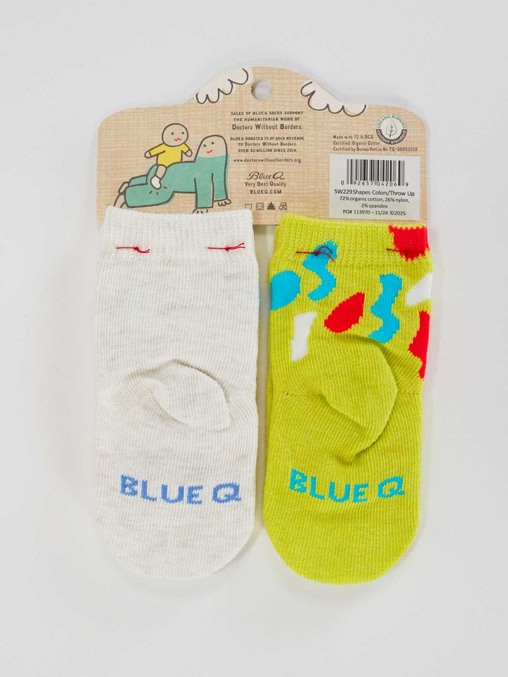 Two pairs of Blue Q baby socks: one white pair with “Cool If I Throw Up On You?” in blue, the other green with colorful shapes inspired by modern art. Fun packaging features cartoon characters. Product names: Big Fan Of Shapes &amp; Colors and Cool If I Throw Up On You?.