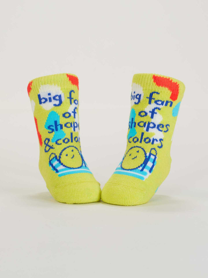 Blue Q’s “Big Fan Of Shapes &amp; Colors” baby socks: yellow modern art design with colorful shapes, a smiling face, and “big fan of shapes &amp; colors” text. Made from soft, OCS-certified organic cotton.