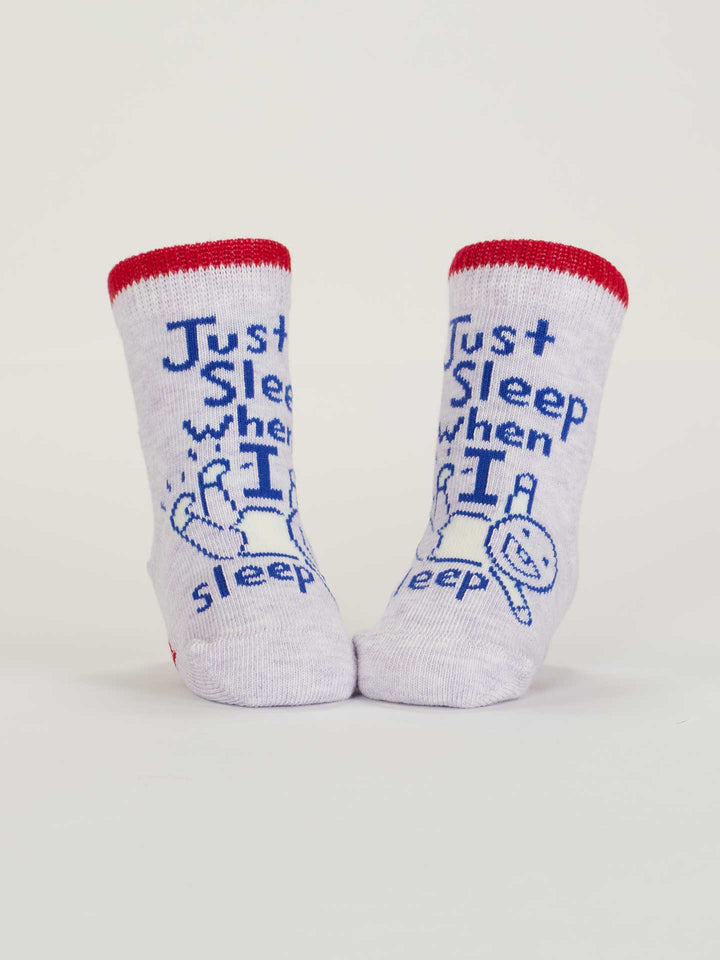 A pair of OCS certified organic cotton Blue Q baby socks, red cuffs, blue "Just Sleep When I Sleep. Lol." text and a sleeping baby drawing on the front. Product name: Wait &