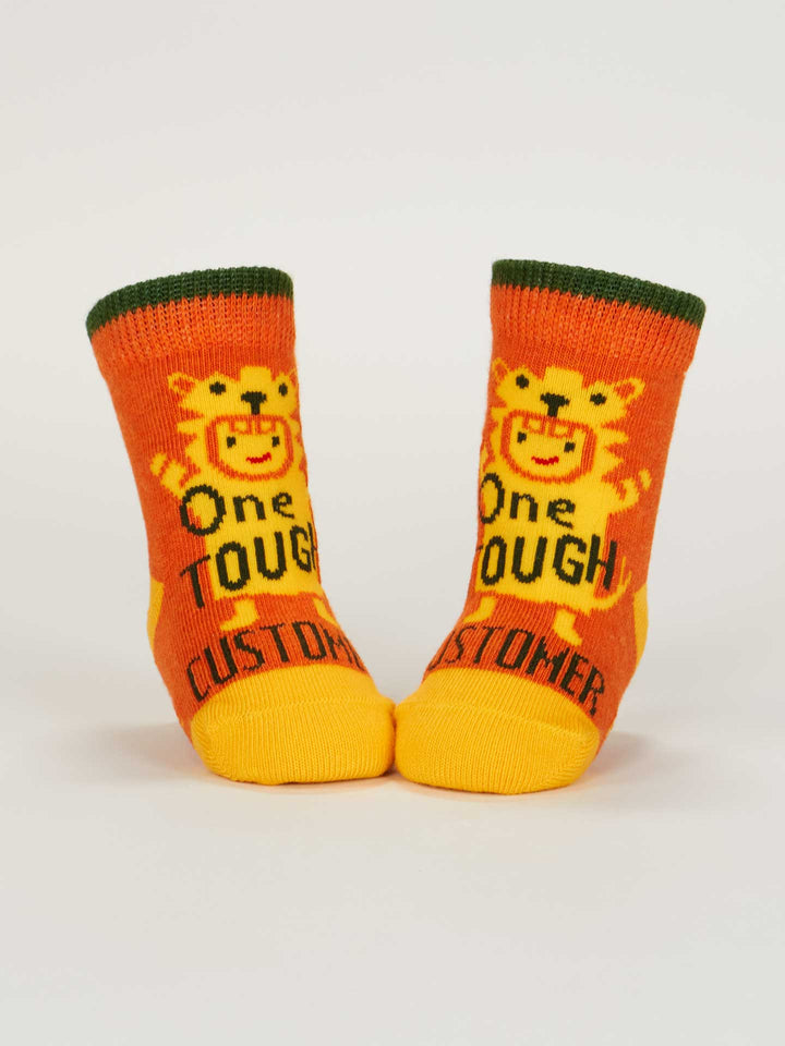 Blue Q’s One Tough Customer/Reflecting On My Behavior Baby Socks (0-12 months) are orange and yellow with green cuffs, showing a cartoon lion and the phrase "One Tough Customer" on the front.