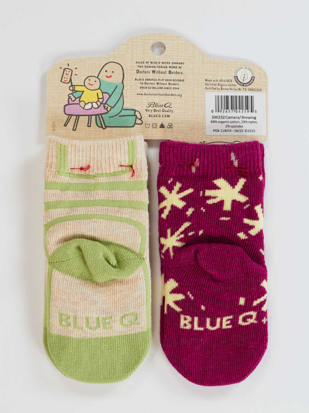 The Camera Loves Me/ I Got One Brewing Baby Socks by Blue Q are cozy 0-12 month socks, attached to a backing. One pair is green with pink stripes and a bear face; the other is magenta with cream stars. "BLUE G." is written near the toes.
