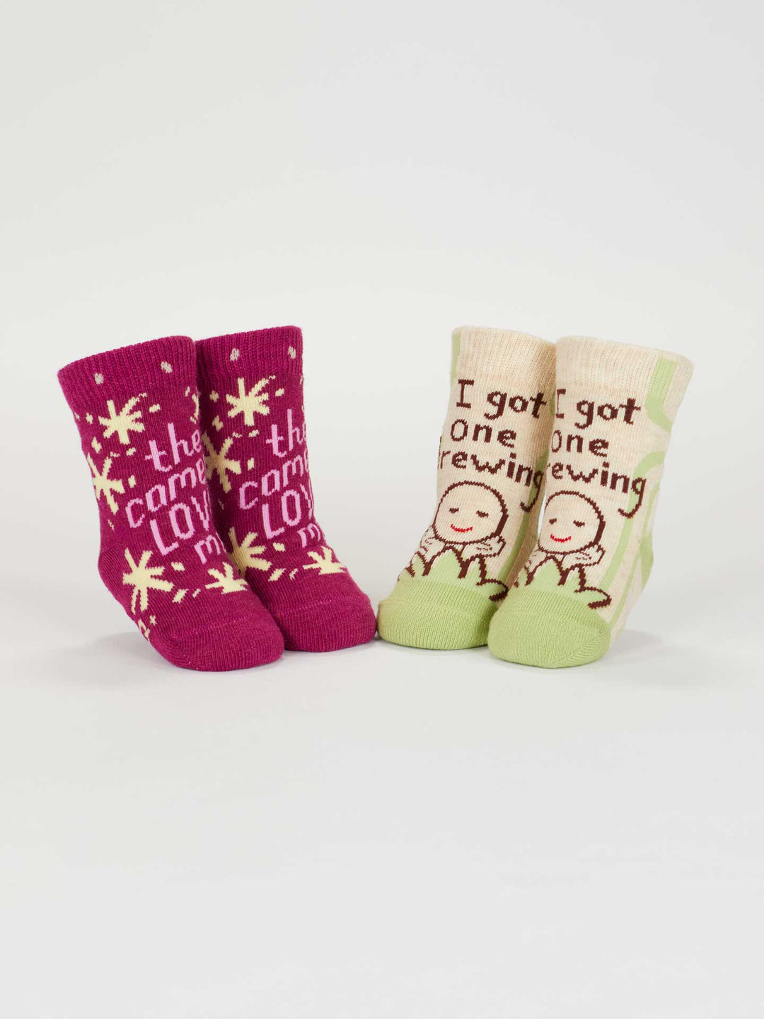 The Blue Q "The Camera Loves Me/I Got One Brewing" baby socks set includes two organic cotton pairs: magenta with stars and fun text, and cream/green with smiling figures. Perfect for newborns 0-12 months.
