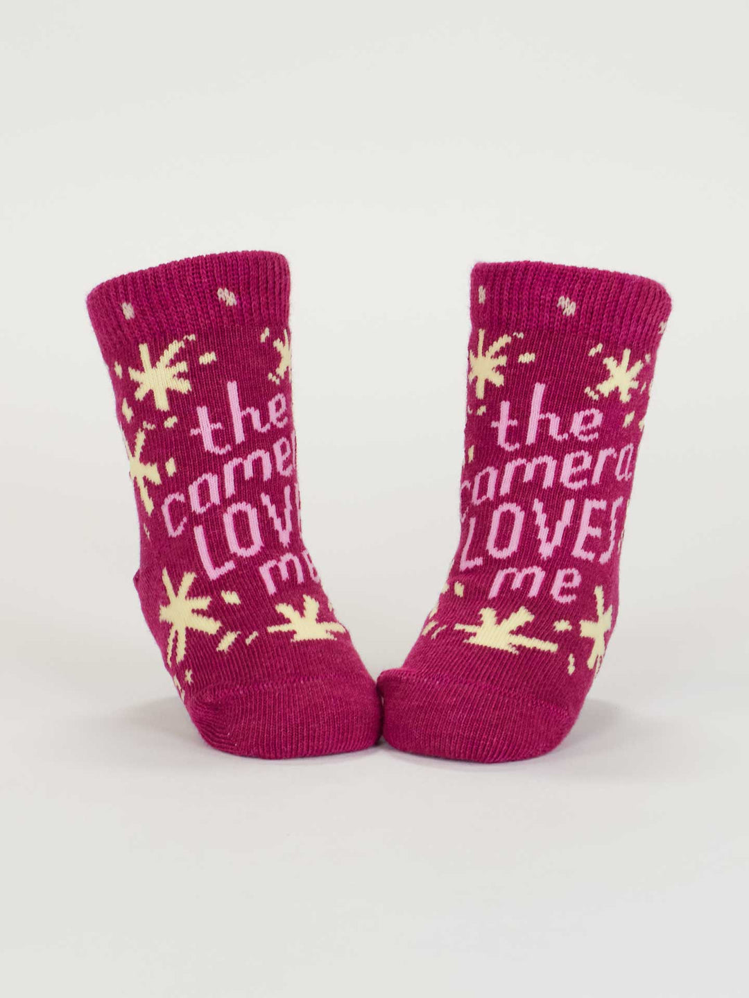 A pair of Blue Q "The Camera Loves Me/ I Got One Brewing" pink organic cotton baby socks, featuring yellow stars and fun text, stand upright against a white background. Ideal for babies aged 0-12 months.