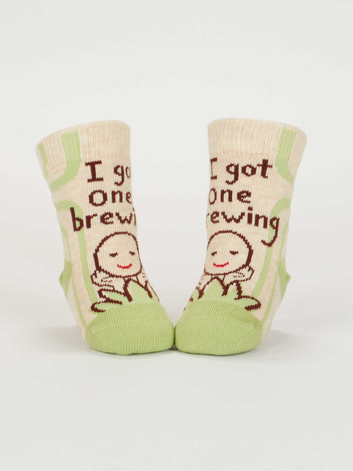 Blue Q’s “The Camera Loves Me/ I Got One Brewing” baby socks feature cream and green organic cotton, a smiling cartoon, and “I got one brewing” print—perfect for babies 0-12 months. Shown standing on a white background.