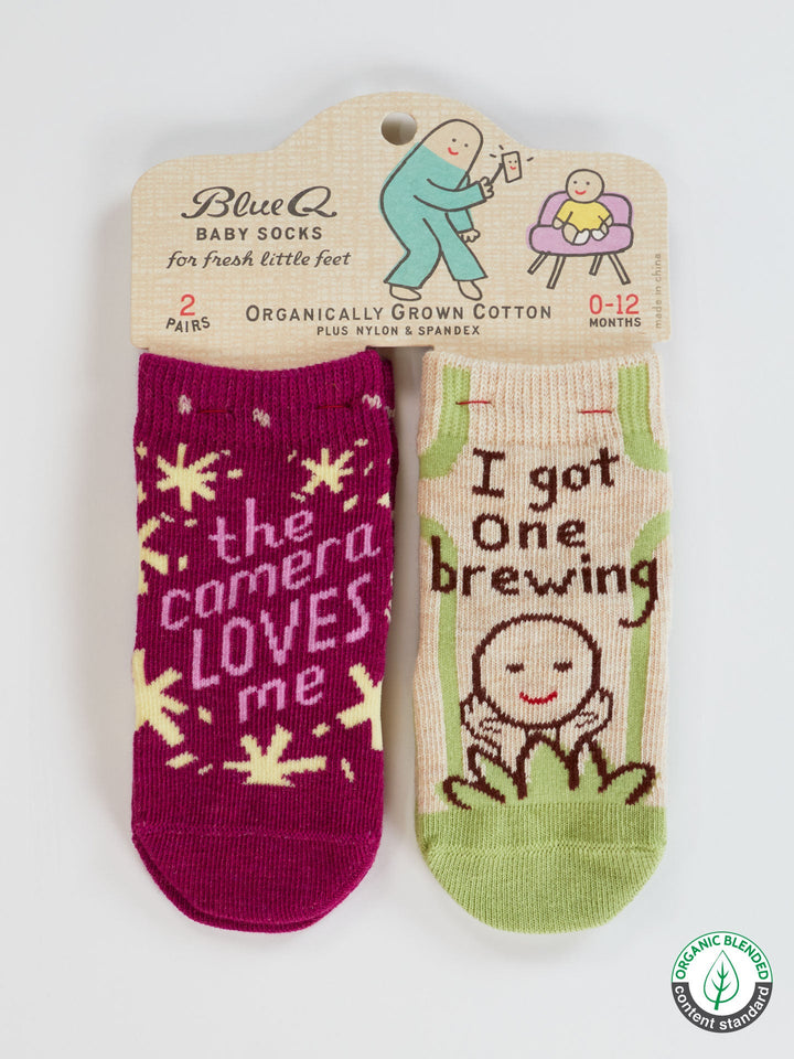 Blue Q’s “The Camera Loves Me/ I Got One Brewing” organic cotton baby socks set includes one magenta pair with yellow stars and one green pair, perfect for newborns ages 0-12 months.