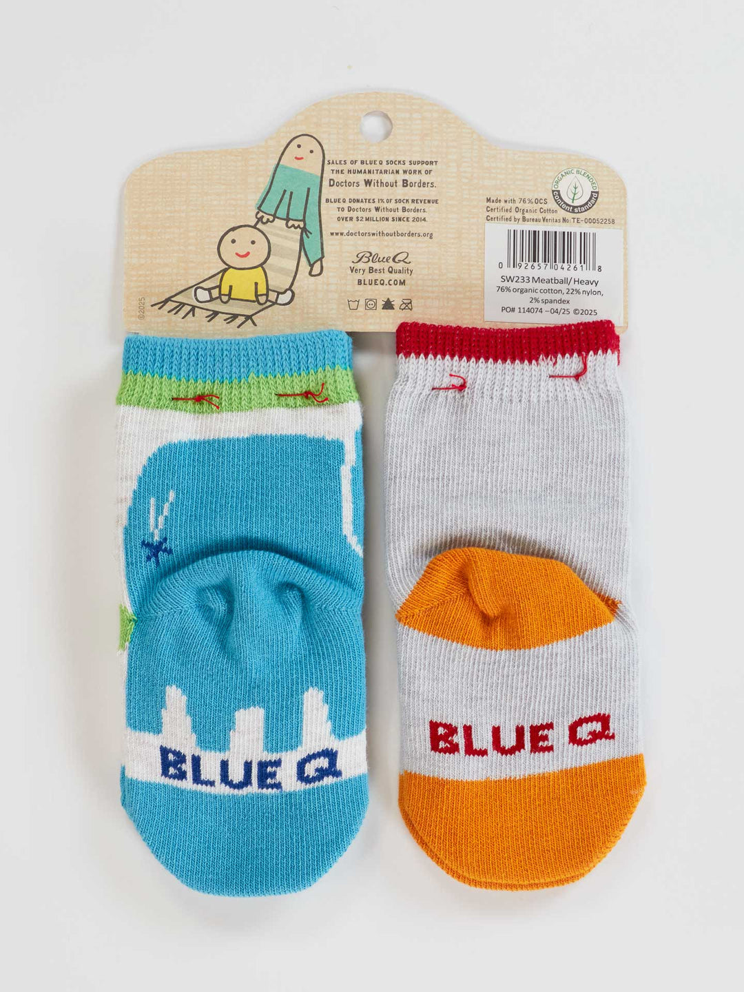 Two pairs of Blue Q baby socks, "Little Meatball" and "I Get Heavy," made from organic cotton. One is blue with a white and green design, the other orange and gray with "BLUE Q" text. Playful hanger features cartoons and product info.