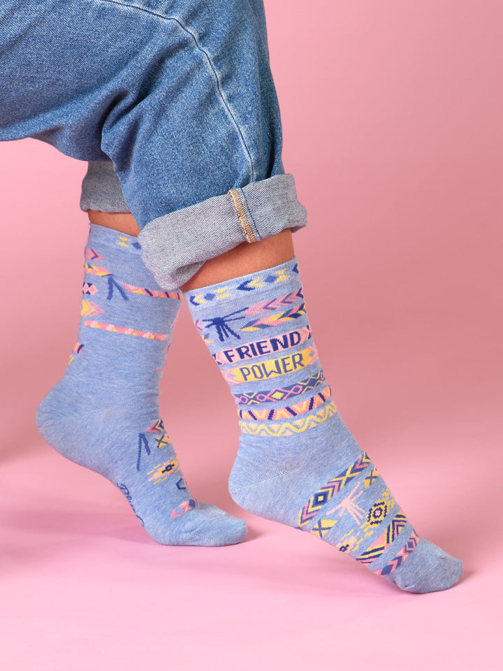 Wearing Blue Q’s Friend Power W-Crew Socks—blue combed cotton with colorful geometric patterns and FRIEND POWER text—styled with rolled-up jeans against a pink backdrop for a sustainable fashion statement.