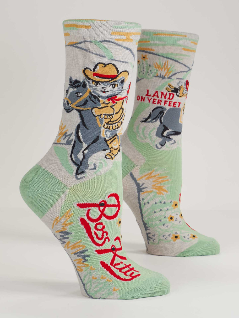 Blue Q’s Boss Kitty W-Crew Socks in light green and gray feature a cartoon cat cowboy on horseback, “Land on yer feet” and “Puss Kitty” text, plus illustrated grass, small cats, and wild frontier accents. Made from combed cotton.
