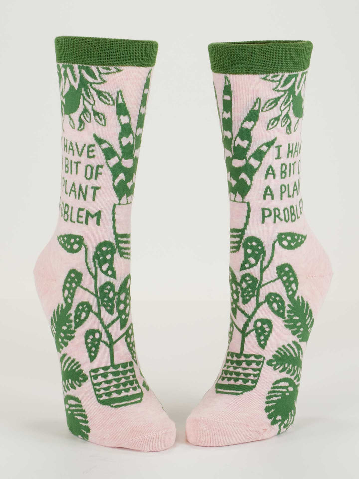 Blue Q’s I Have A Bit Of A Plant Problem W-Crew Socks are pink women’s socks with green trim, made from soft combed cotton. They feature the phrase "I HAVE A BIT OF A PLANT PROBLEM" plus monstera leaves and potted plants for a playful look.
