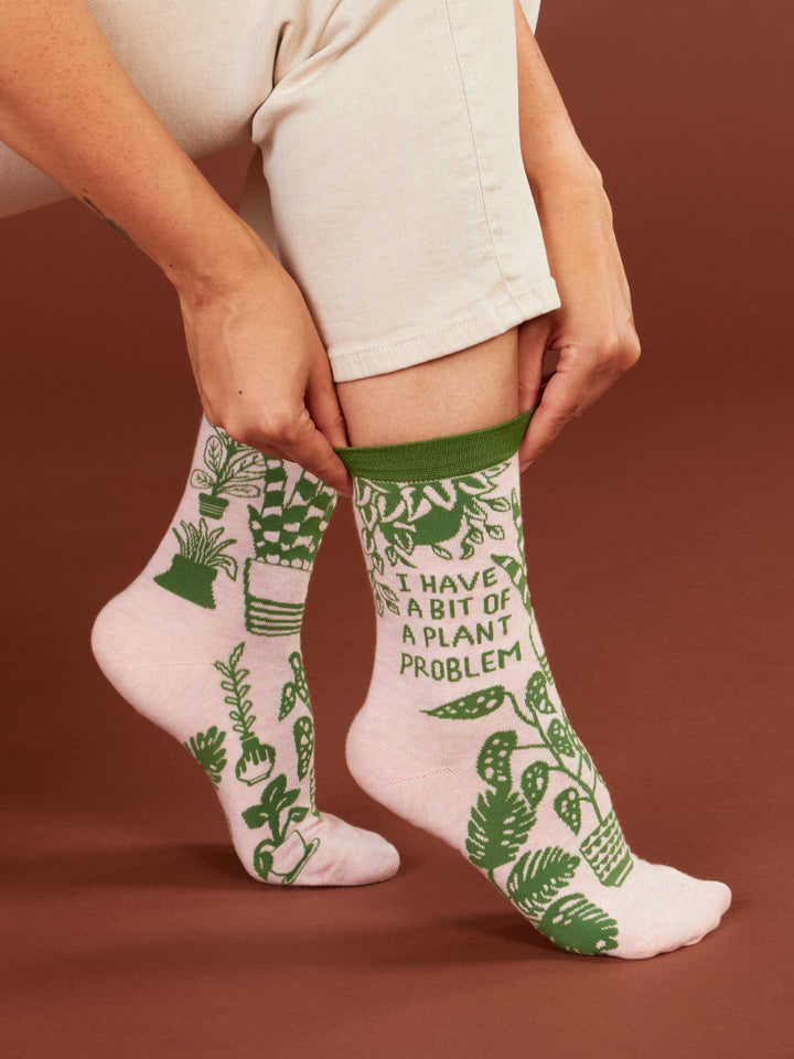 A person adjusts a Blue Q I Have A Bit Of A Plant Problem W-Crew Sock—light pink combed cotton with green monstera designs and text—sitting on a brown background, ideal for pairing with your favorite women&