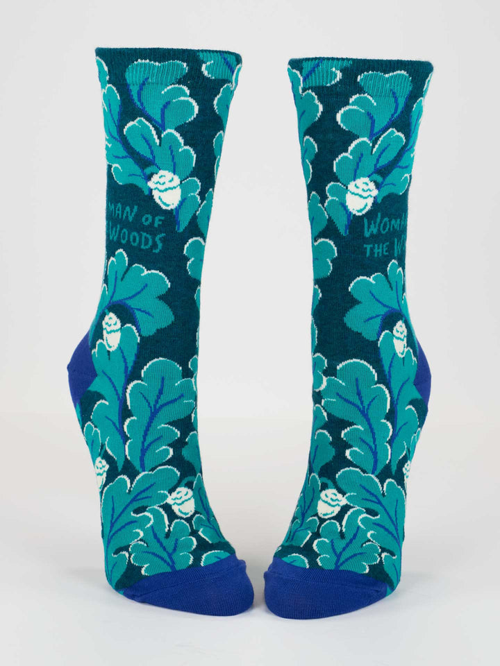 Blue Q Woman Of The Woods W-Crew Socks: teal and blue cotton socks for women, featuring leafy patterns, a hidden figure in the foliage, and “WOMAN OF THE WOODS” text on each sock.