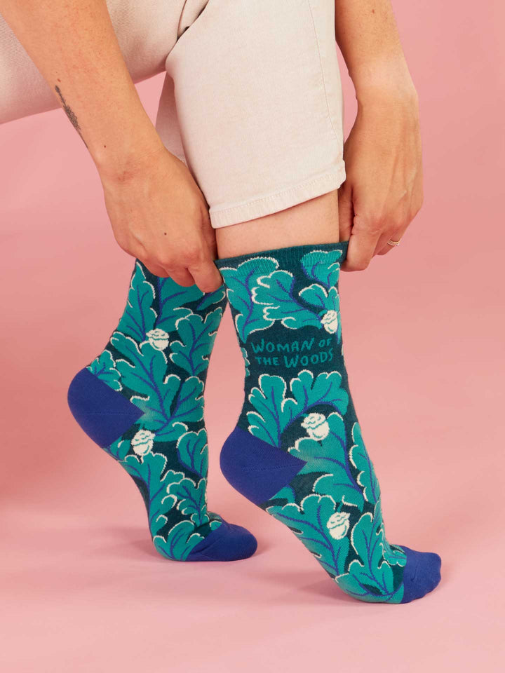 Someone in white pants puts on Blue Q’s Woman Of The Woods W-Crew Socks—teal cotton socks featuring leafy patterns, white faces, and "WOMAN OF THE WOODS" text on a pink background.