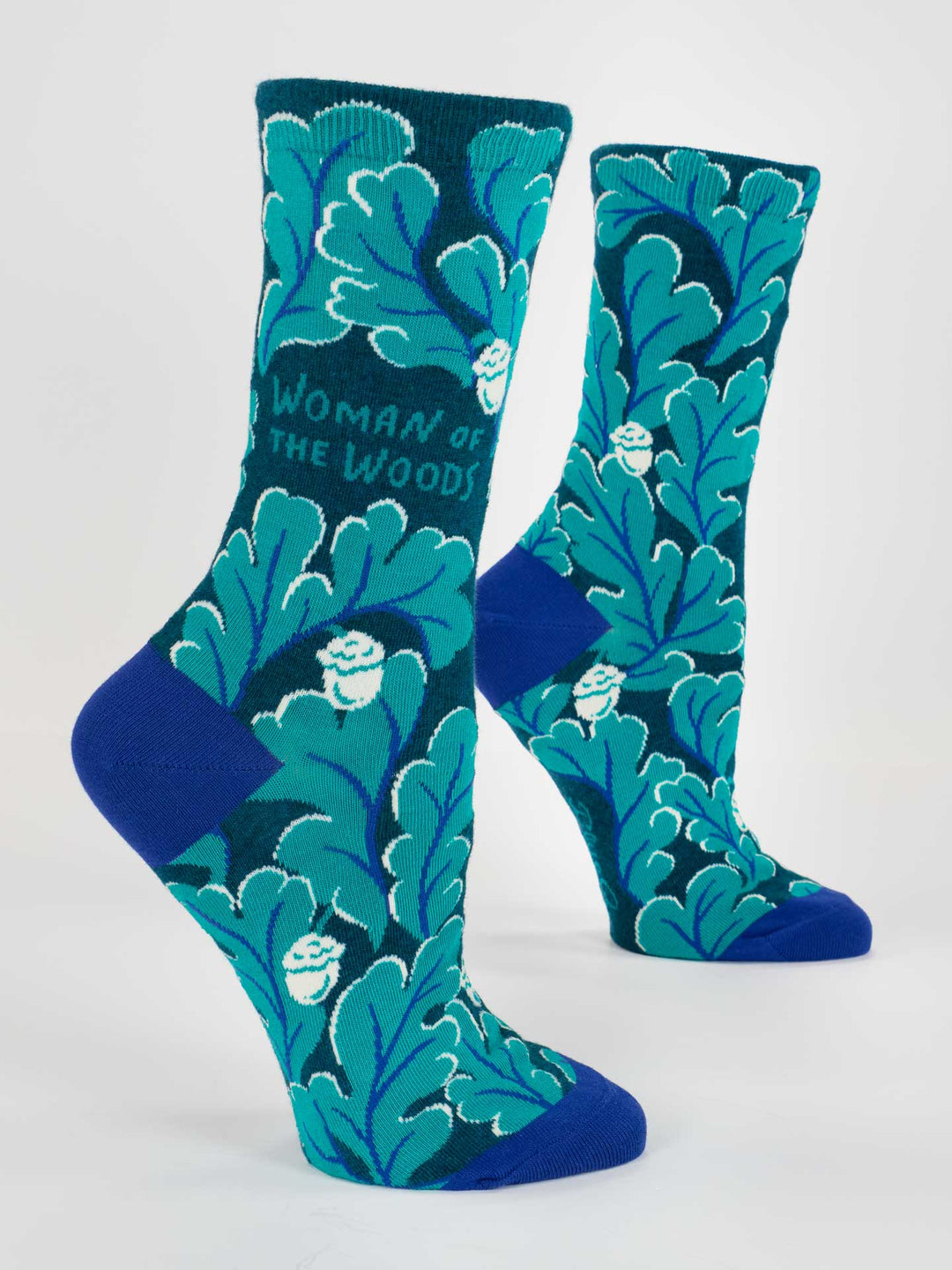 Blue Q's Woman Of The Woods W-Crew Socks are blue and teal women's socks featuring leafy patterns and "Woman of the Woods" text, shown on mannequin feet against a white background—ideal cotton socks for nature lovers.