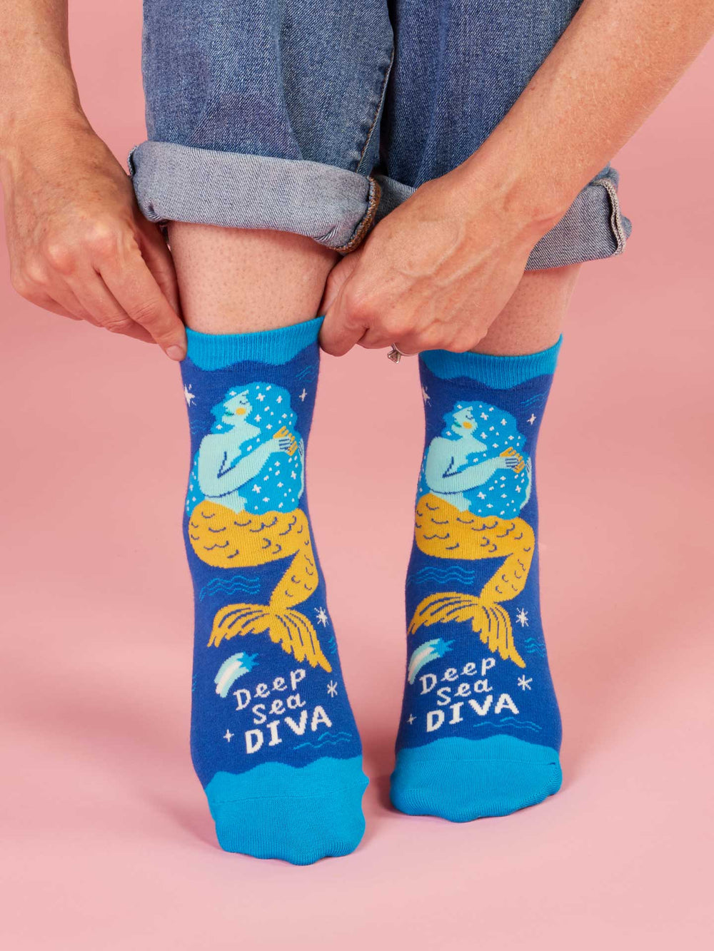 A person with rolled-up jeans adjusts bright blue Blue Q Deep Sea Diva W-Ankle Socks, featuring a yellow-tailed mermaid, sea stars, and “Deep Sea Diva” text on pink. These playful women’s socks add fun to any outfit.