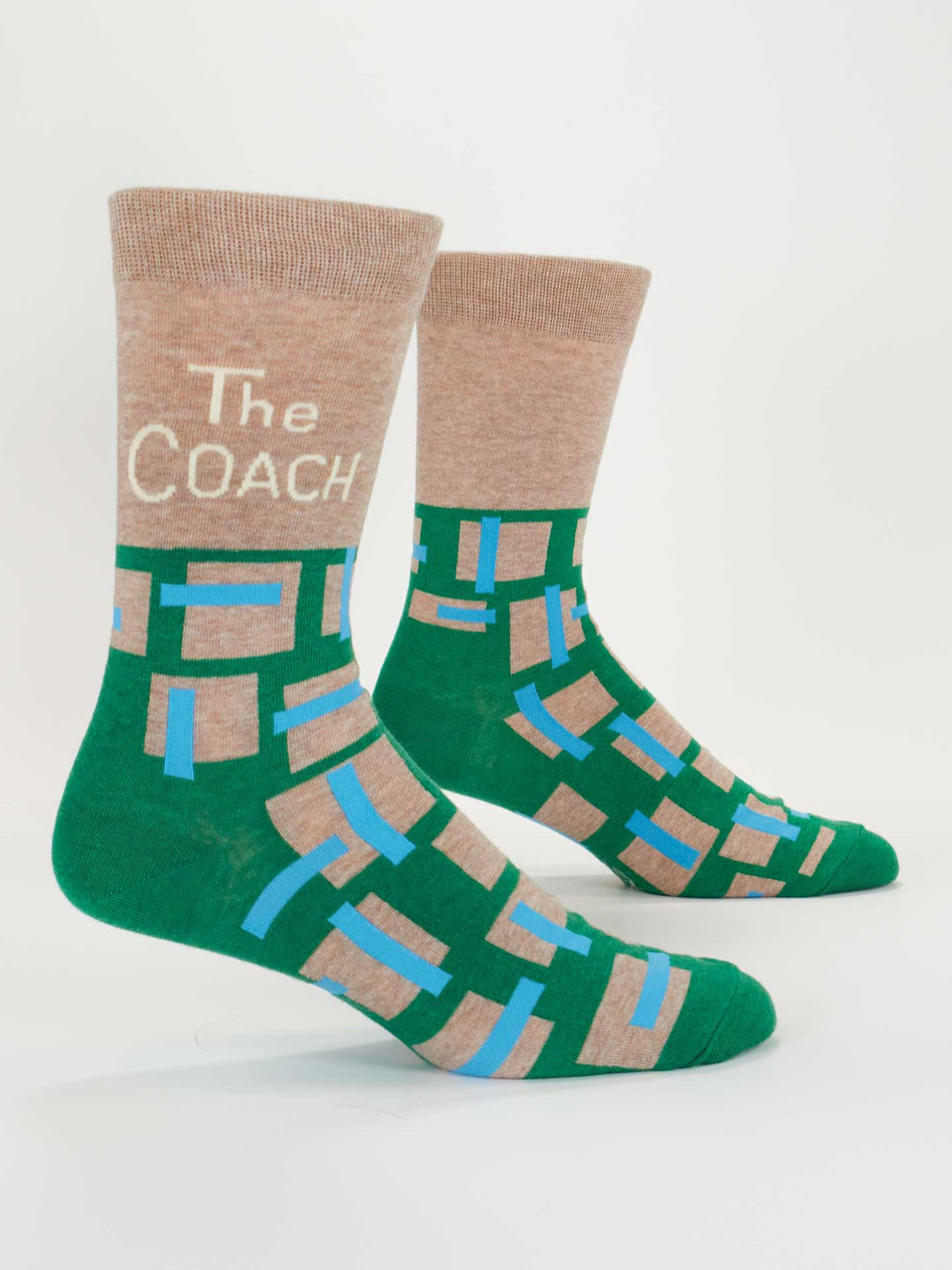 The Coach M-Crew Socks by Blue Q: two green combed cotton men's socks with beige toes, heels, and cuffs. Each cuff has "COACH" in white and features a geometric beige and blue pattern on foot and ankle.
