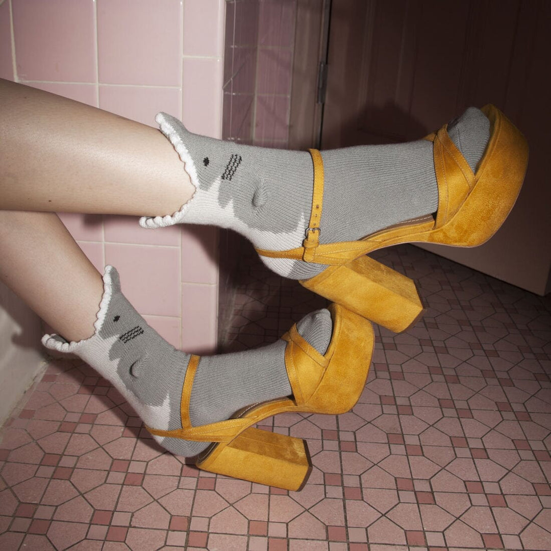 Model wearing the grey 3D shark socks with yellow platform heels on pink tile