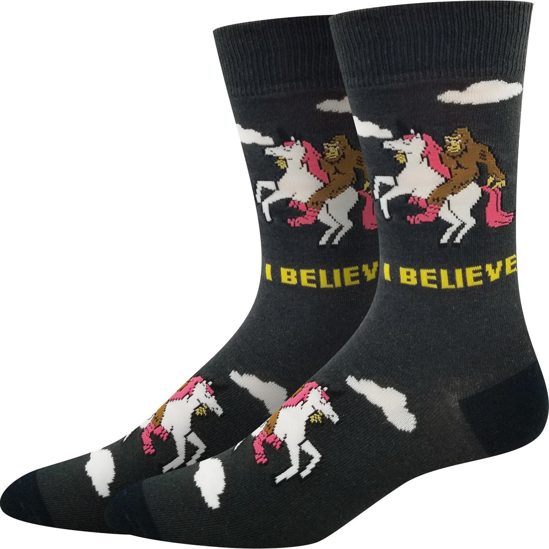 Pair of charcoal unisex crew socks showing Bigfoot riding a pink unicorn with I Believe lettering and clouds