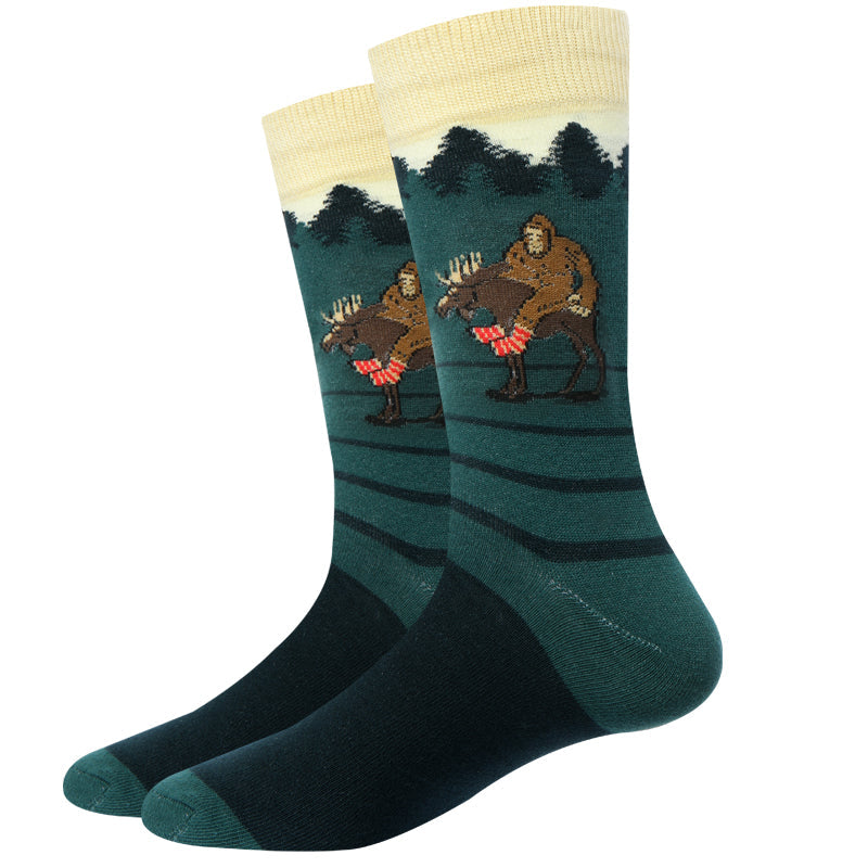 Pair of green Moose Bigfoot crew socks displayed flat, with a cream cuff and a Sasquatch riding a moose through striped evergreens