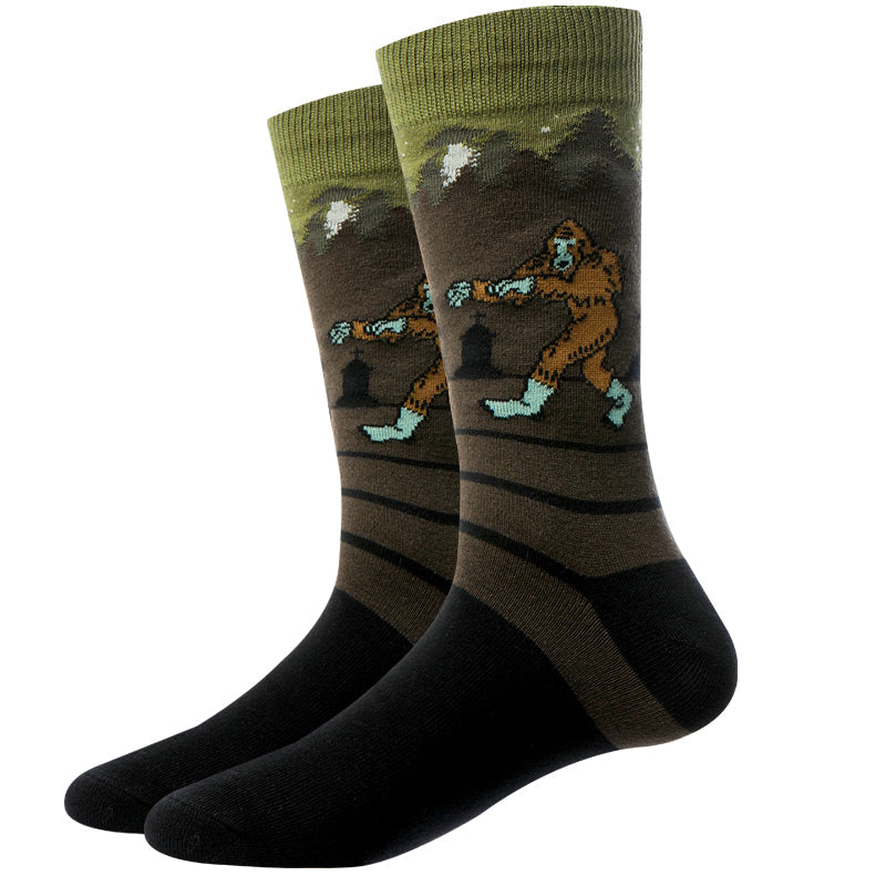 Pair of olive and brown Zombie Bigfoot crew socks displayed flat, with a zombie Sasquatch and mint-green limbs in a moonlit forest