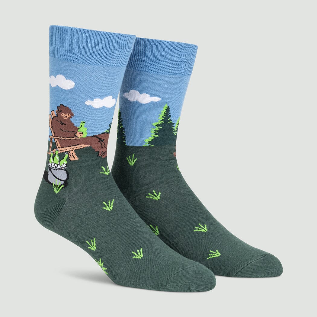 Men's crew socks with Bigfoot relaxing in a camp chair with a drink in a forest meadow, blue cuff and green grassy foot, on foot forms