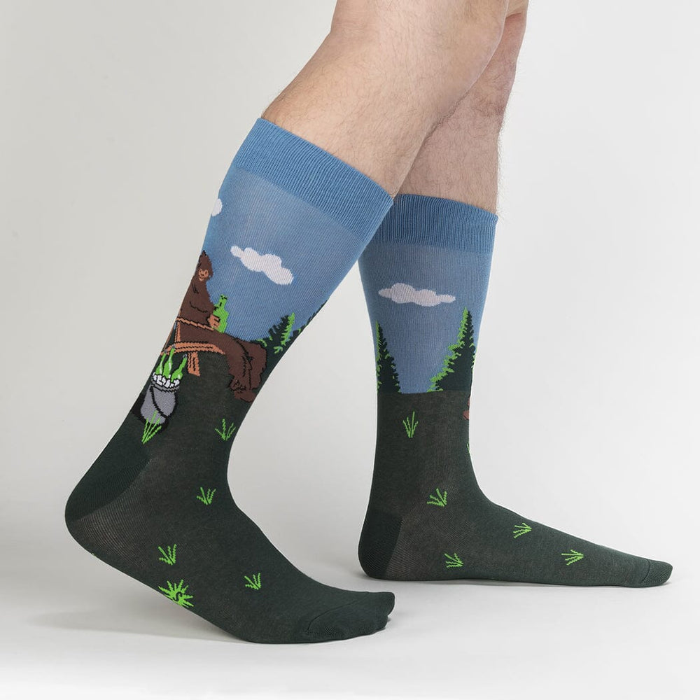 Bigfoot Bucket List socks worn on a model's legs, side view showing Sasquatch in the chair