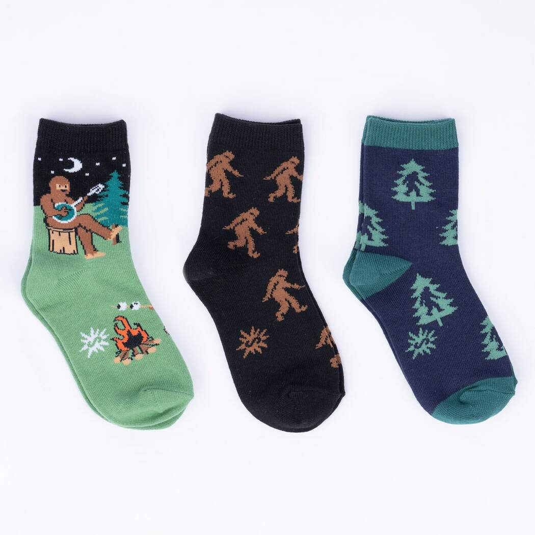 Three kids' camping crew socks flat: Bigfoot banjo campfire pair, brown Sasquatch print pair, and navy pine tree pair