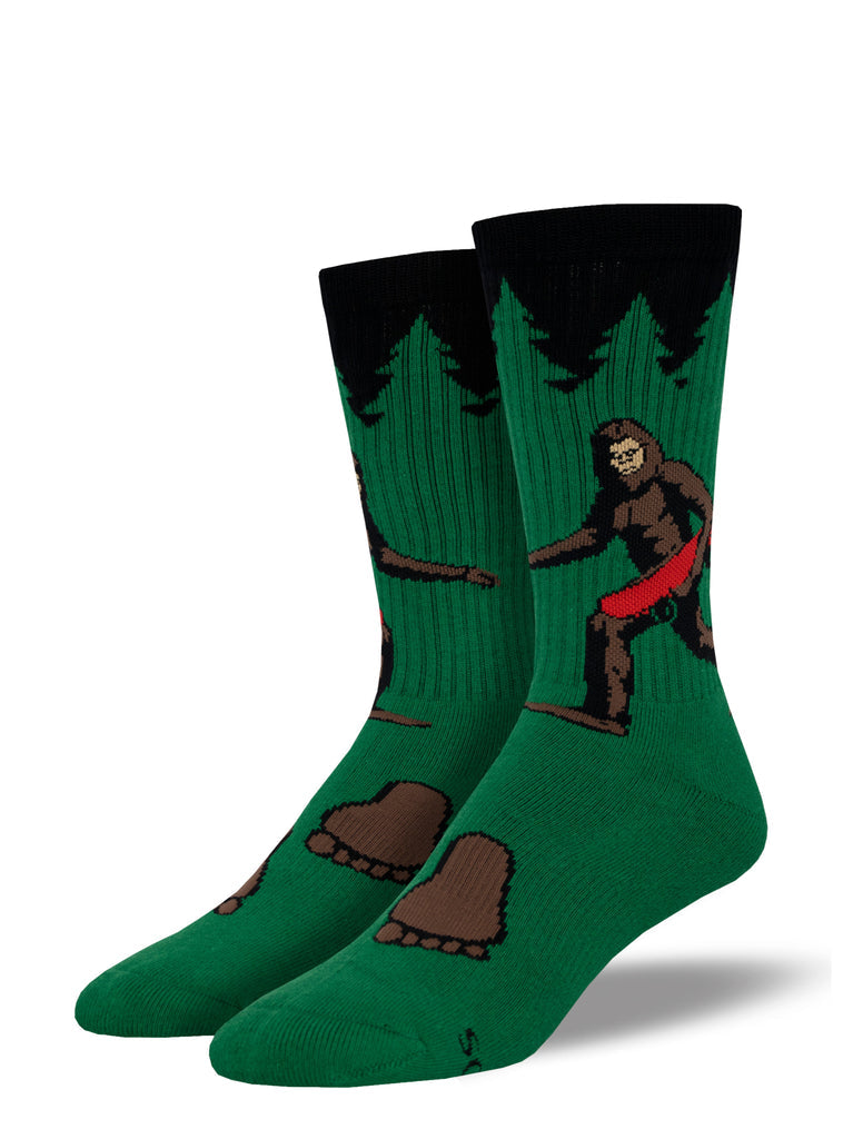 Green men's athletic crew socks with black cuff showing a brown Bigfoot riding a skateboard among evergreens, with footprints on the foot