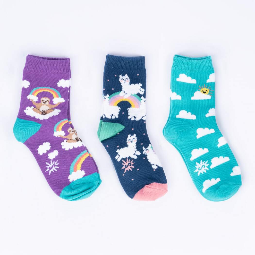 Flat-lay of three kids' crew socks: a purple pair with sloths on rainbows, a navy pair with alpacas, and a teal pair with clouds and a sun