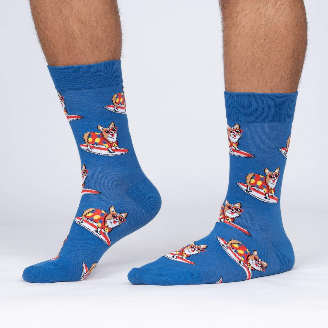 Men's blue crew socks worn on a model's legs, showing corgis in sunglasses and tropical shirts surfing on red boards