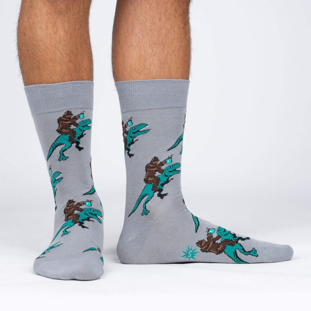 Men's gray crew socks worn on a model's legs, showing Bigfoot holding coffee while riding a teal T-rex