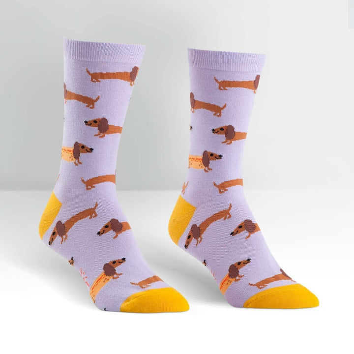 Pair of lavender crew socks covered in dachshunds, some dressed as hot dogs, with yellow heel and toe