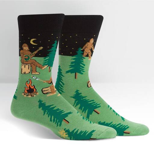 Pair of green Sasquatch campout crew socks shown flat, one playing banjo by a fire among pine trees