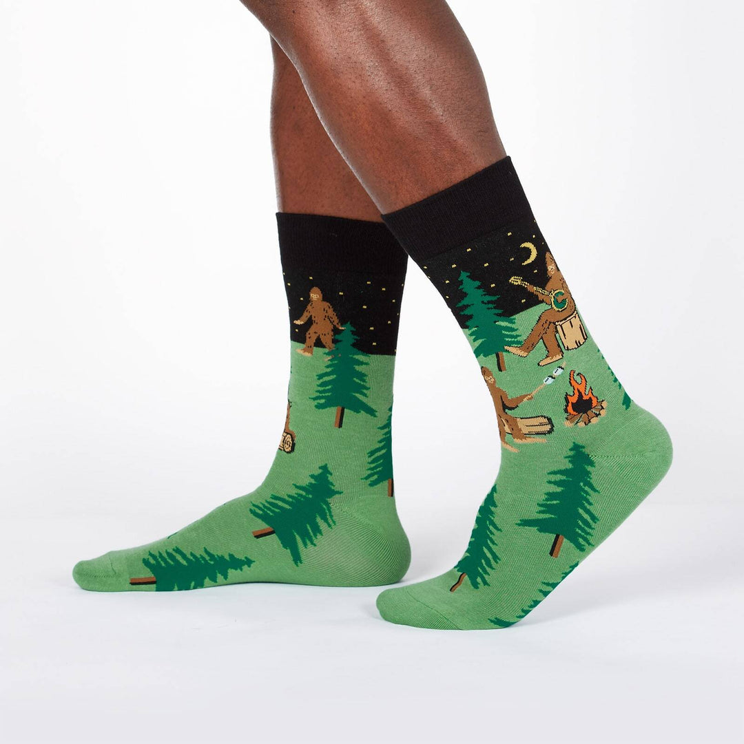 Green Bigfoot camping crew socks worn on a model's legs, showing Sasquatches around a campfire under a starry night cuff