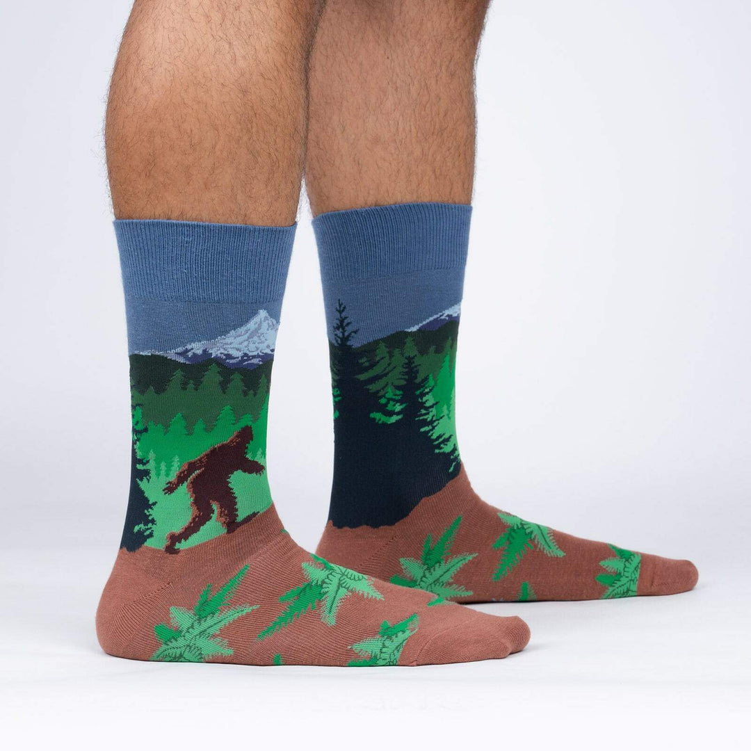 Men's crew socks worn on a model's legs, showing a Bigfoot silhouette in a green forest below snowy mountains, rust-brown feet